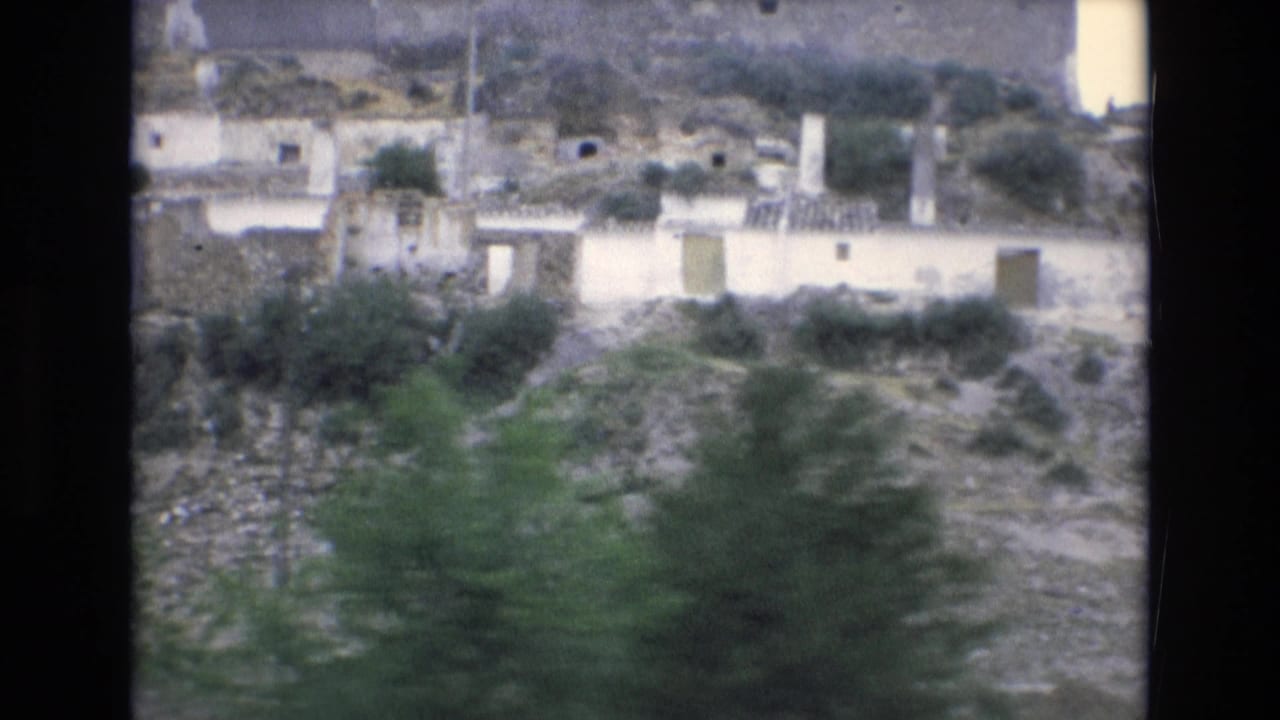 Rocky hillside settlement with pale buildings, trees, shrubs, and an upper stone structure.