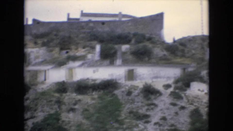 The 1972 Spain footage moves closer to low whitewashed hillside buildings, showing dirt paths, uneven rocky ground, shrubs, and small door and window openings.