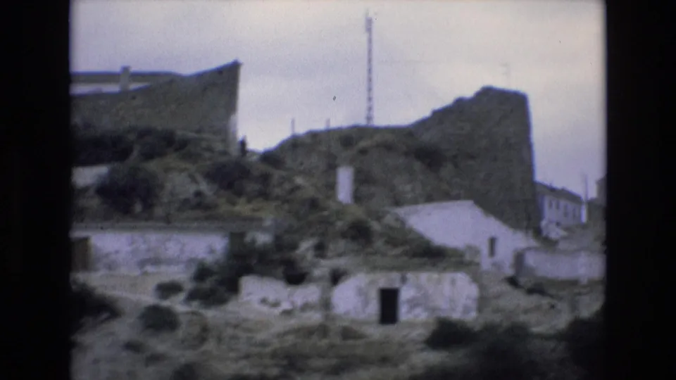 A soft 8mm frame from Spain in 1972 shows the edge of a hillside village with small pale buildings, a dark doorway, stone walls, trees, and a rocky ridge.