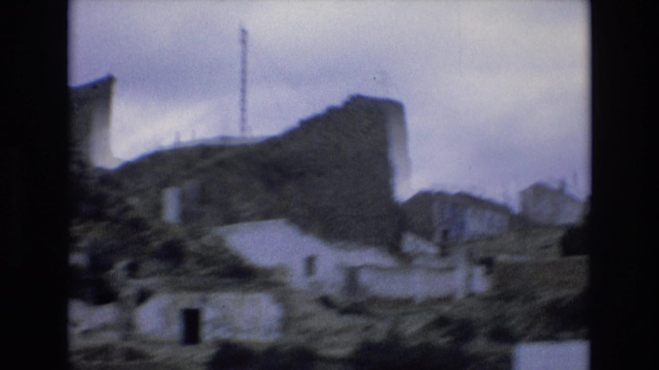 Soft 8mm frame from Spain in 1972 showing whitewashed hillside structures, rough stonework, scrubby slopes, and an overcast sky from a passing vehicle.
