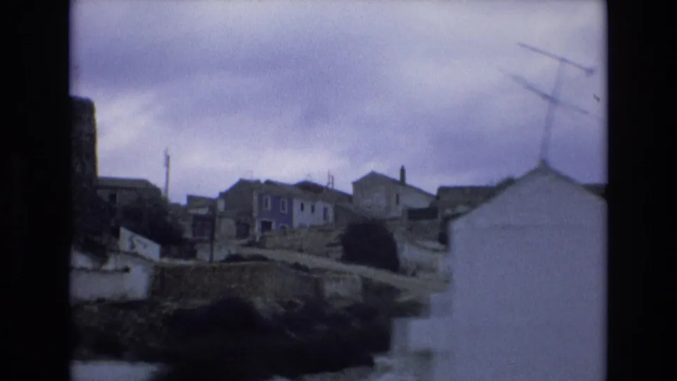 1972 Spain home-movie view of village houses along a hillside road, with stone walls, white building exteriors, and a distant mast under an overcast sky.