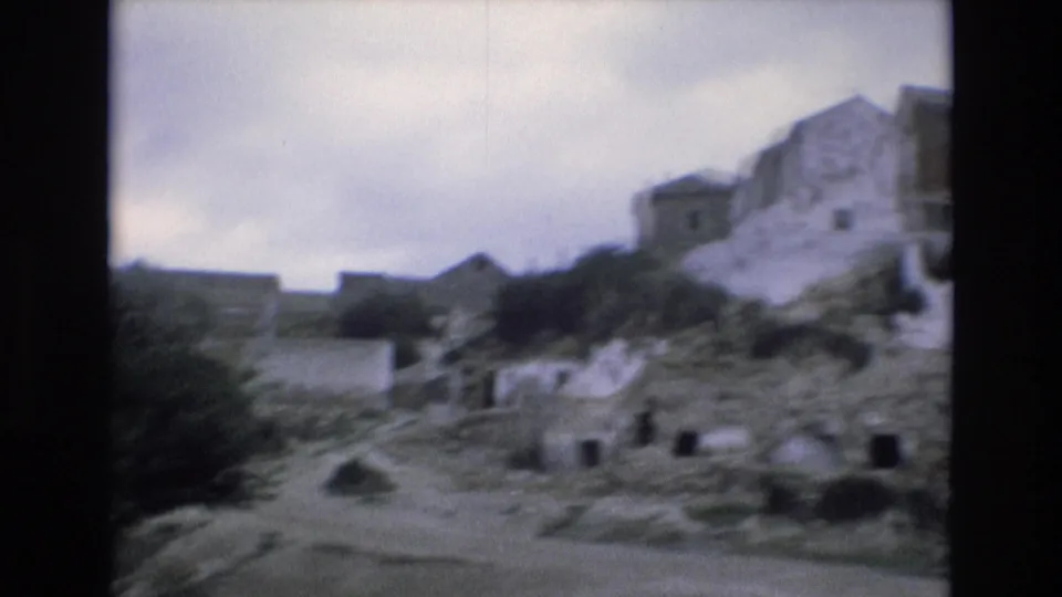 Moving 8mm view in Spain from 1972 showing a rocky village slope, whitewashed buildings, rough ground, small dark openings, and low vegetation.