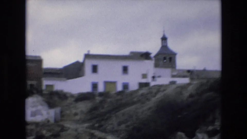 1972 Spain 8mm frame showing a white village building and church bell tower set into a grassy hillside beneath a cloudy sky.
