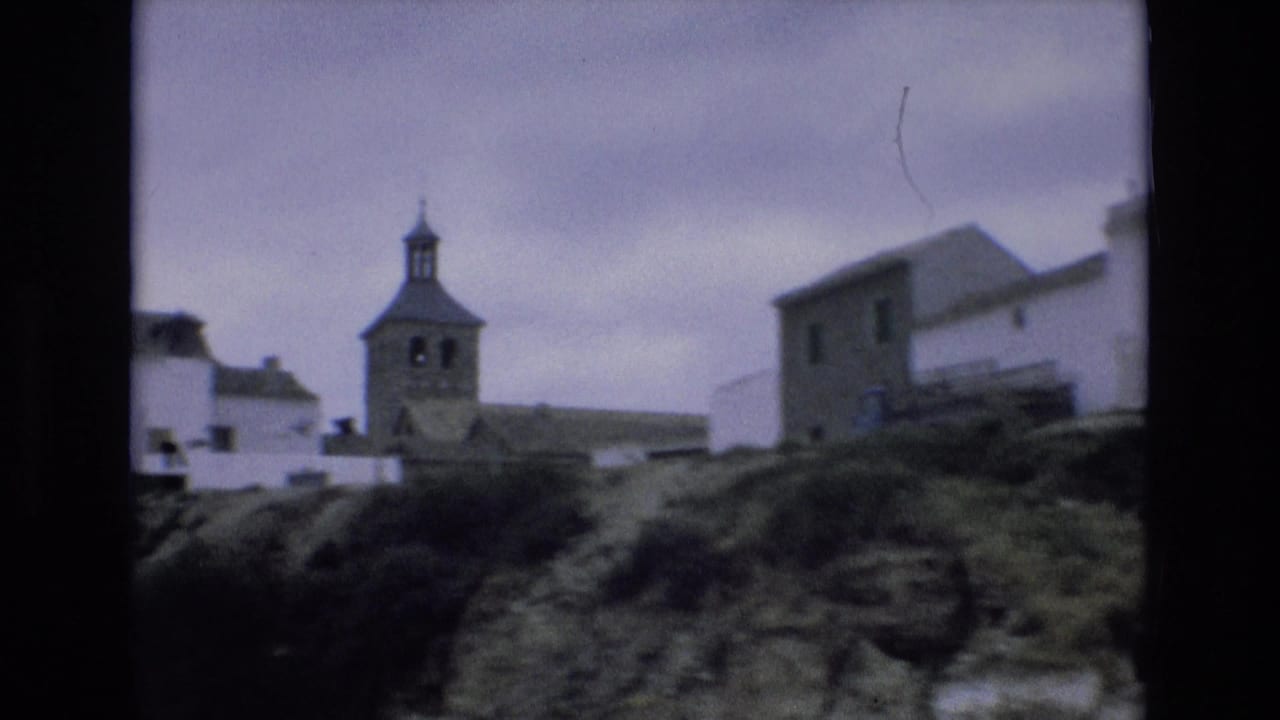 1972 Spain home-movie frame showing a hillside village with white buildings, a church bell tower, rough ground, and overcast sky.