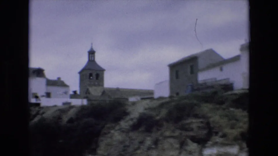 1972 Spain home-movie frame showing a hillside village with white buildings, a church bell tower, rough ground, and overcast sky.