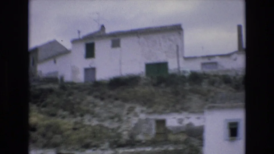 1972 Spain travel frame showing whitewashed structures on a slope, including a building with a green door or gate and rocky foreground.