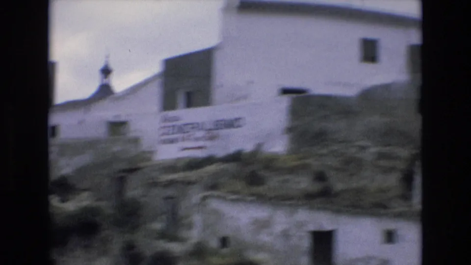 1972 Spain frame with a blurred painted wall sign referencing Generalisimo, white village buildings, hillside terrain, and a church dome behind the structures.