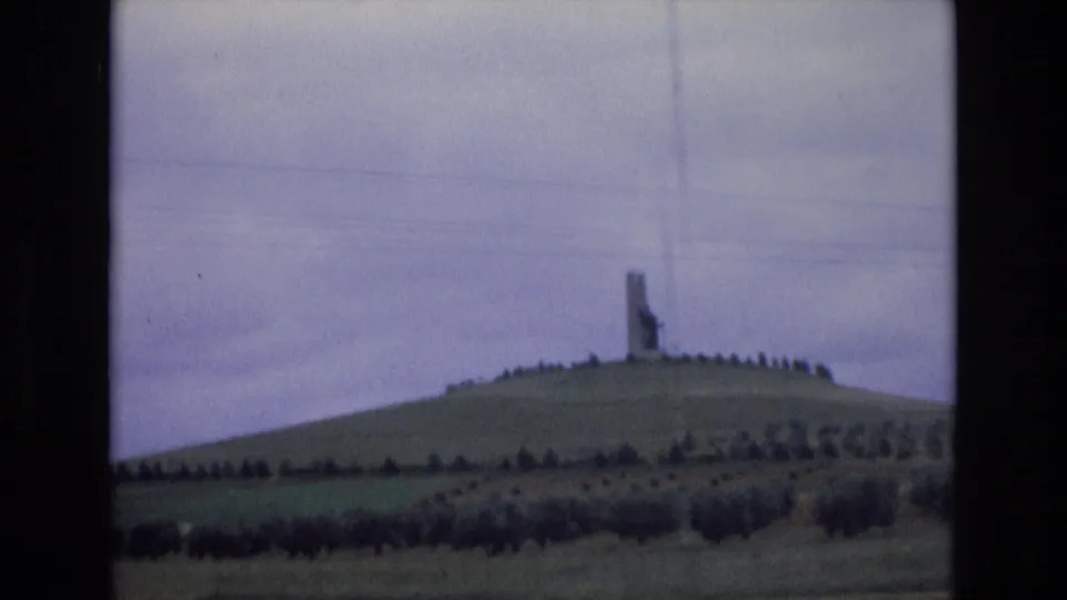 1972 Spain home-movie frame showing a distant hilltop monument or statue, grassy slopes, cultivated fields, tree line, and overcast sky.