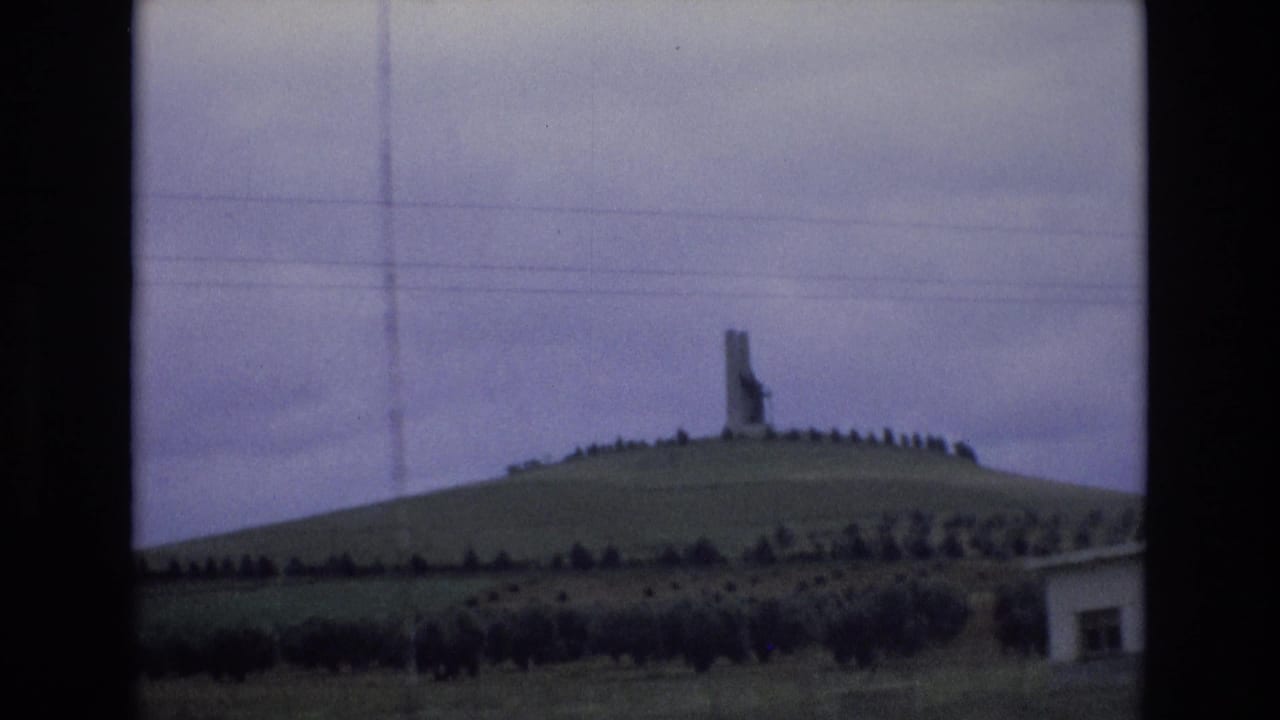 1972 Spain 8mm frame showing a tall hilltop monument or marker above a ring of trees or shrubs, with overcast sky and utility wires crossing the view.