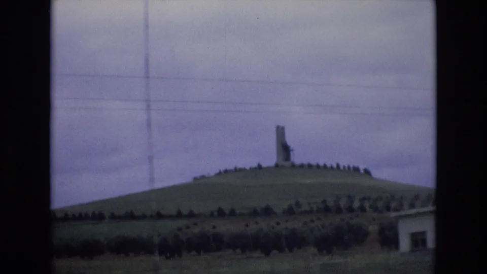 1972 Spain 8mm frame showing a tall hilltop monument or marker above a ring of trees or shrubs, with overcast sky and utility wires crossing the view.
