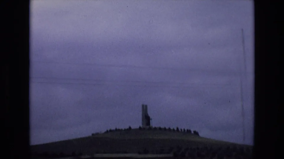 Distant 1972 Spain view of a slender monument on a grassy summit, framed by a line of trees or shrubs and crossed by overhead utility wires.