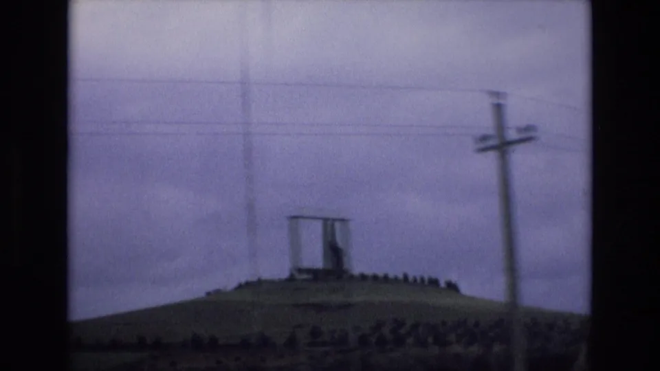 1972 Spain home-movie frame with a utility pole in the foreground, wires across the sky, and a distant hilltop monument behind a circular line of trees or shrubs.