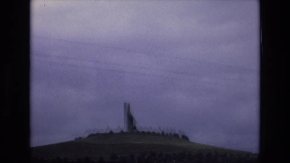 Soft 8mm frame from 1972 Spain showing a distant hilltop monument or landmark, a low ring of trees or shrubs, darker vegetation below, and an overcast sky.