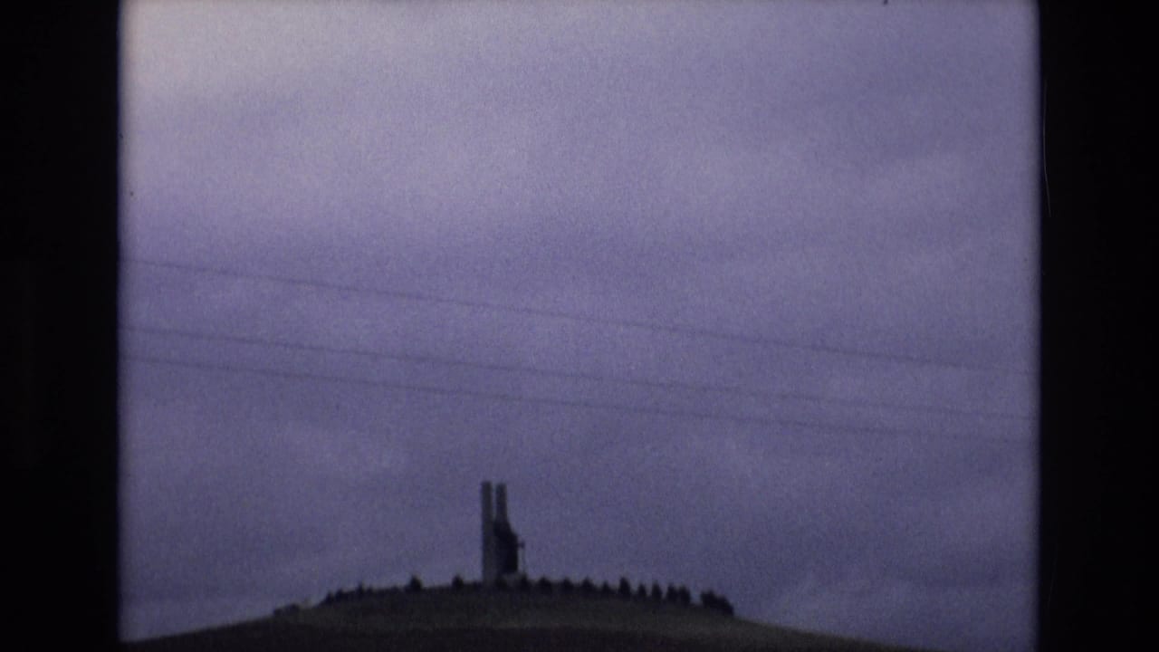 Spain 1972 8mm frame showing a distant tall structure on a grassy hilltop beneath purple-gray clouds, with overhead utility lines crossing the view.