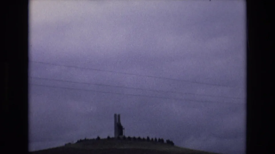 Spain 1972 8mm frame showing a distant tall structure on a grassy hilltop beneath purple-gray clouds, with overhead utility lines crossing the view.