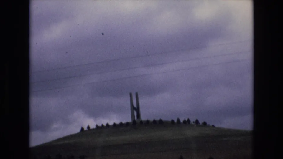 Spain 1972 8mm frame showing the tall summit structure partly framed by a ring of small rocks or trees, with muted landscape tones and overhead wires visible against the clouds.