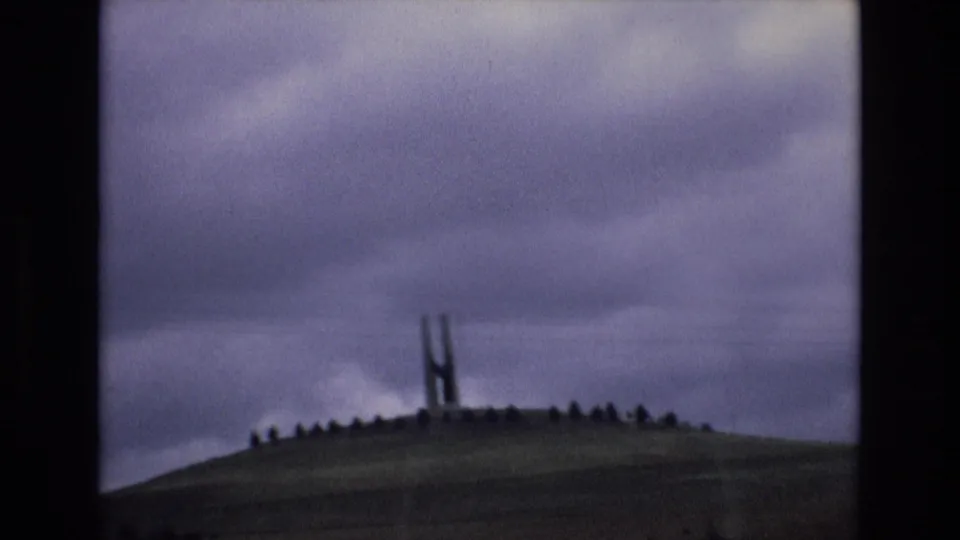 Spain 1972 8mm frame holding on the hilltop structure and ridge markers beneath a broad overcast sky, with the soft focus and dark borders of scanned home-movie film.