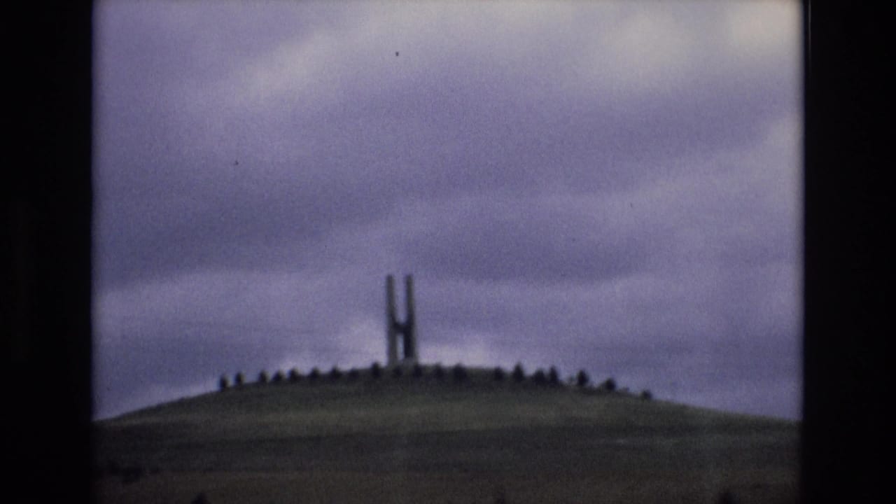 1972 Spain 8mm frame showing a distant H-shaped hilltop monument on a rounded grassy mound under an overcast purplish-gray sky.