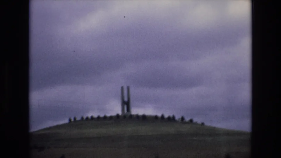 1972 Spain 8mm frame showing a distant H-shaped hilltop monument on a rounded grassy mound under an overcast purplish-gray sky.