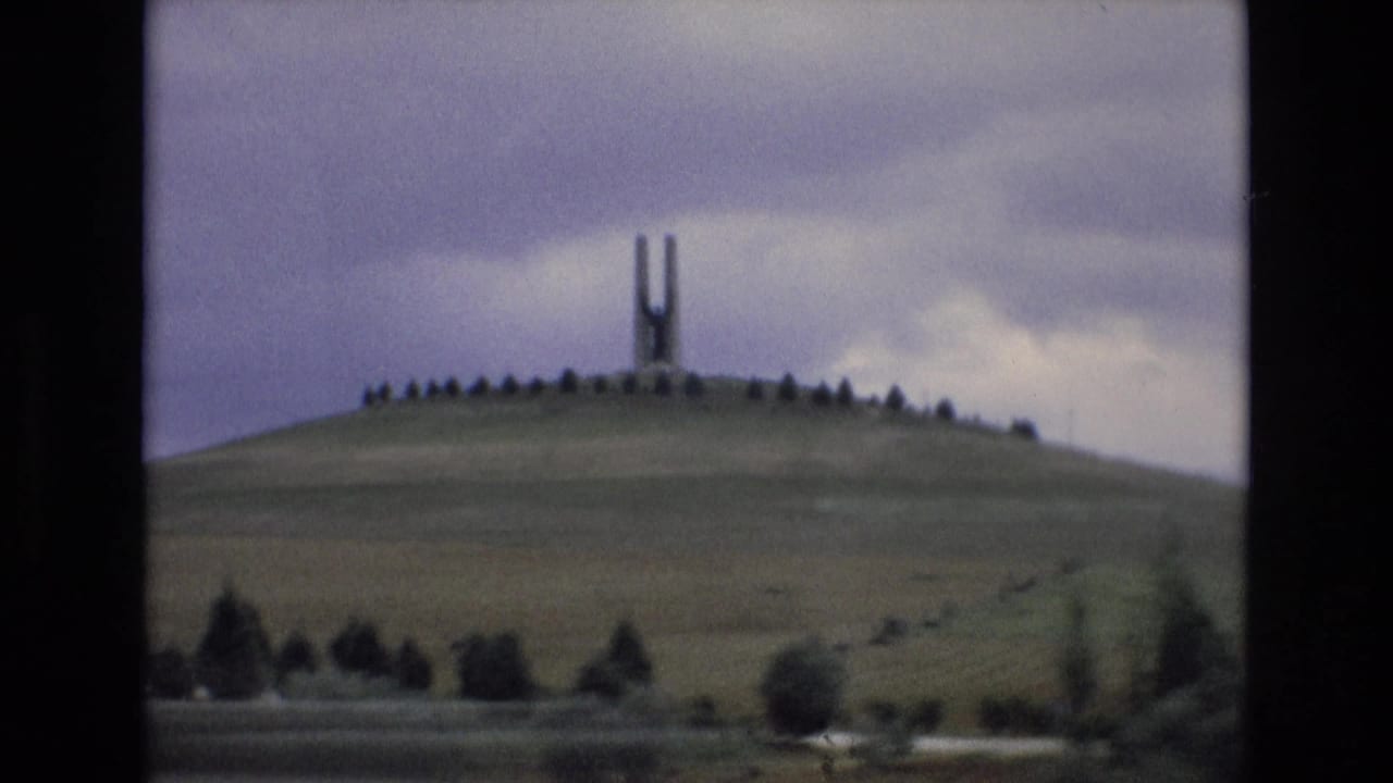 Hilltop H-shaped monument surrounded by small trees, with open fields below.