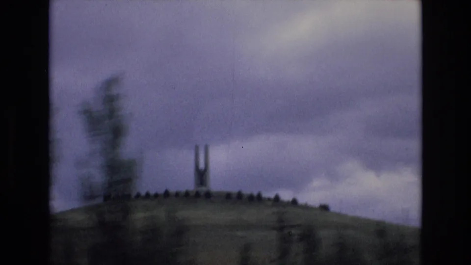 1972 Spain 8mm frame with motion blur from a passing foreground tree partially obscuring the distant H-shaped monument on the hill.