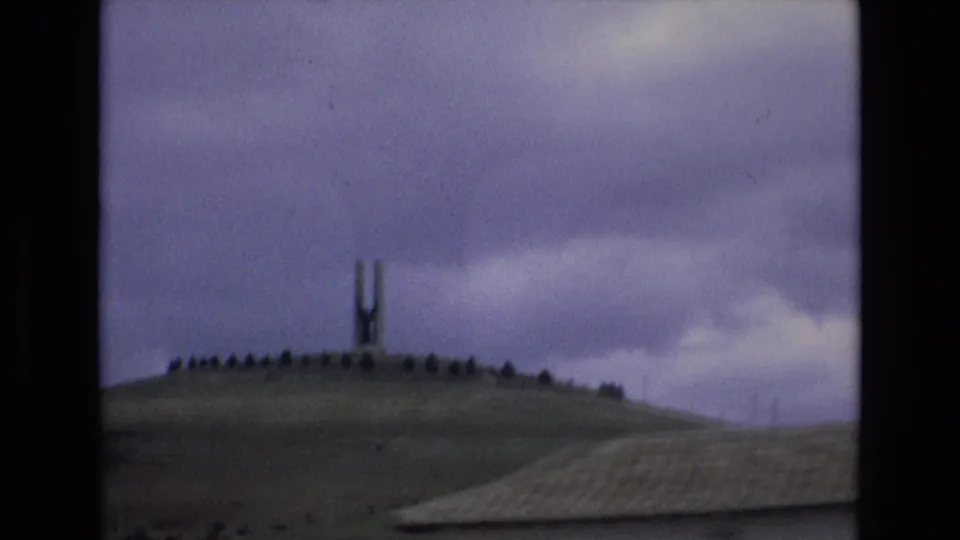 1972 Spain 8mm frame showing the H-shaped monument on a grassy hill, with a sloped roof edge crossing the lower foreground under cloudy sky.