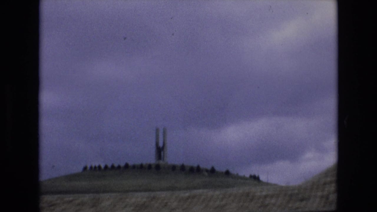 Spain 1972 8mm frame showing a low hillside with an unidentified tall slender structure silhouetted under a cloudy pale purple sky.