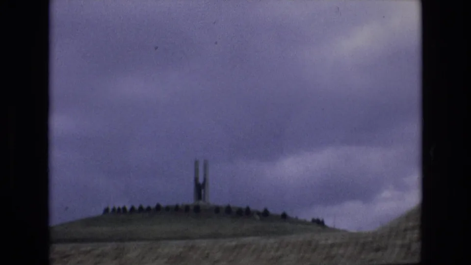 Spain 1972 8mm frame showing a low hillside with an unidentified tall slender structure silhouetted under a cloudy pale purple sky.