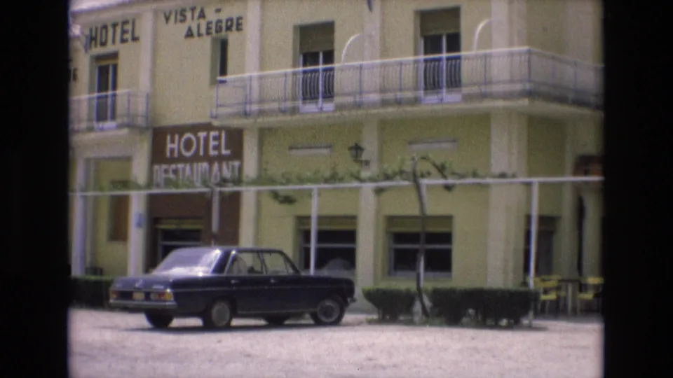 Spain 1972 8mm frame showing the Hotel Vista Alegre frontage, restaurant sign, balconies, ground-floor entrance, hedges, and a dark parked car.