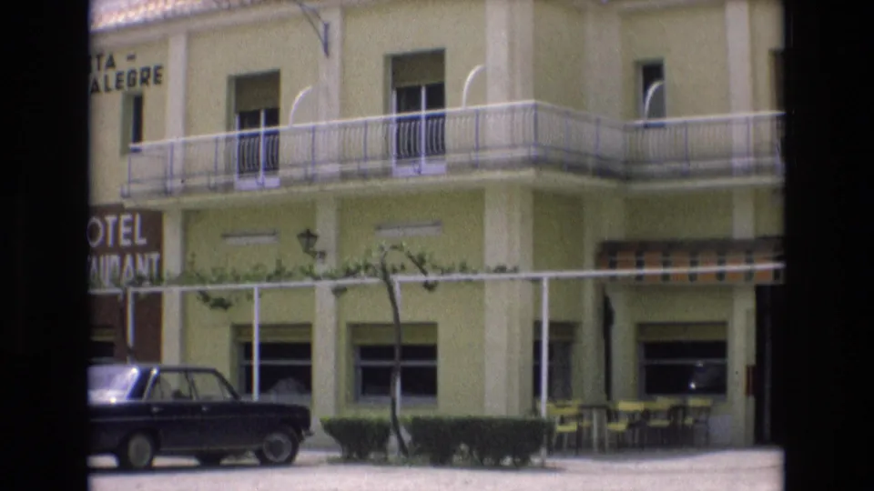 Spain 1972 8mm frame showing part of the Hotel Vista Alegre exterior with balcony railings, restaurant sign edge, trimmed hedges, outdoor tables, and a parked car.
