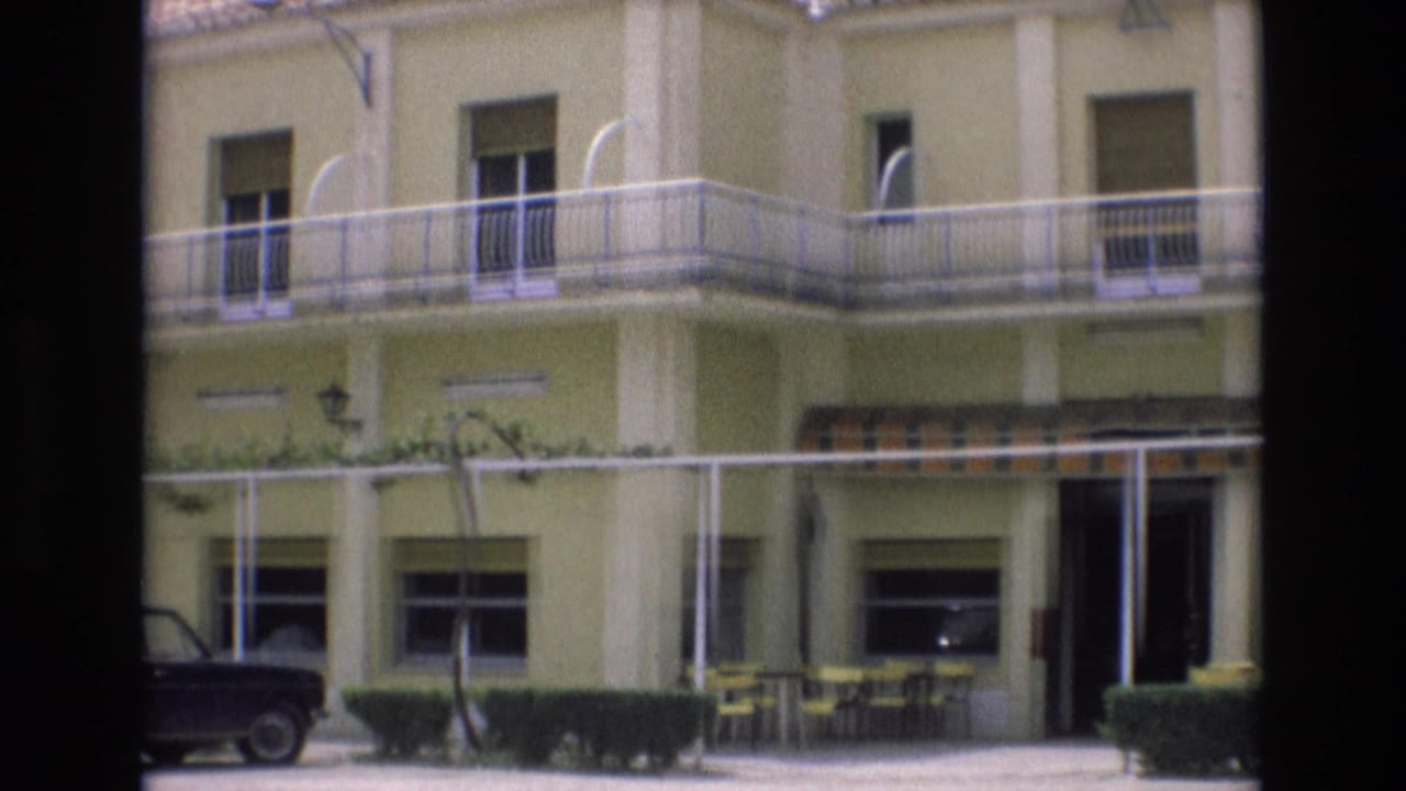 1972 Spain home-movie frame showing a pale yellow motel exterior with white trim, balcony railings, patio furniture beneath a striped awning, hedges, and a dark parked sedan.