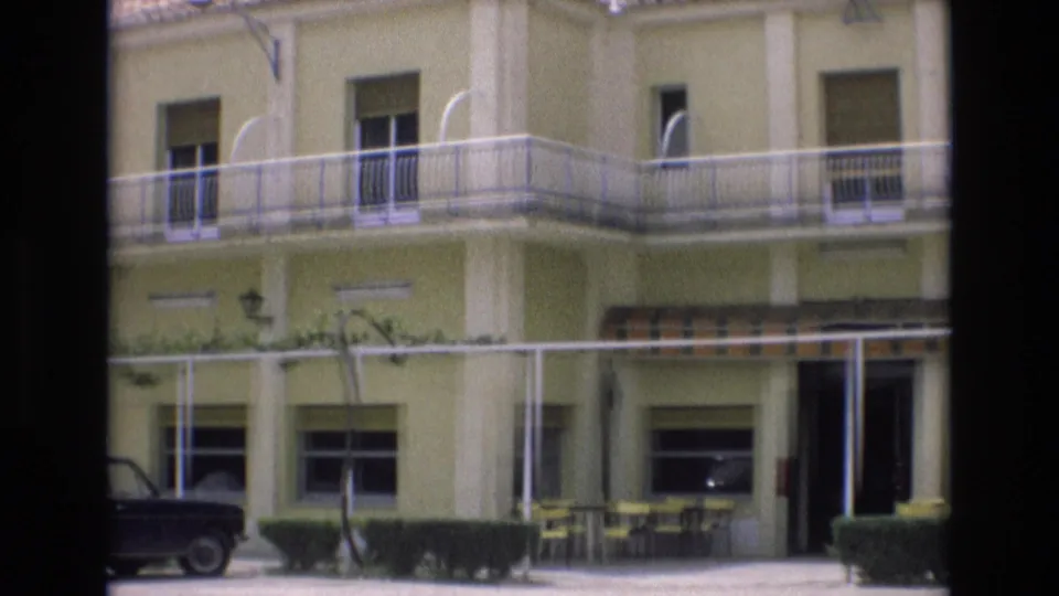 1972 Spain home-movie frame showing a pale yellow motel exterior with white trim, balcony railings, patio furniture beneath a striped awning, hedges, and a dark parked sedan.