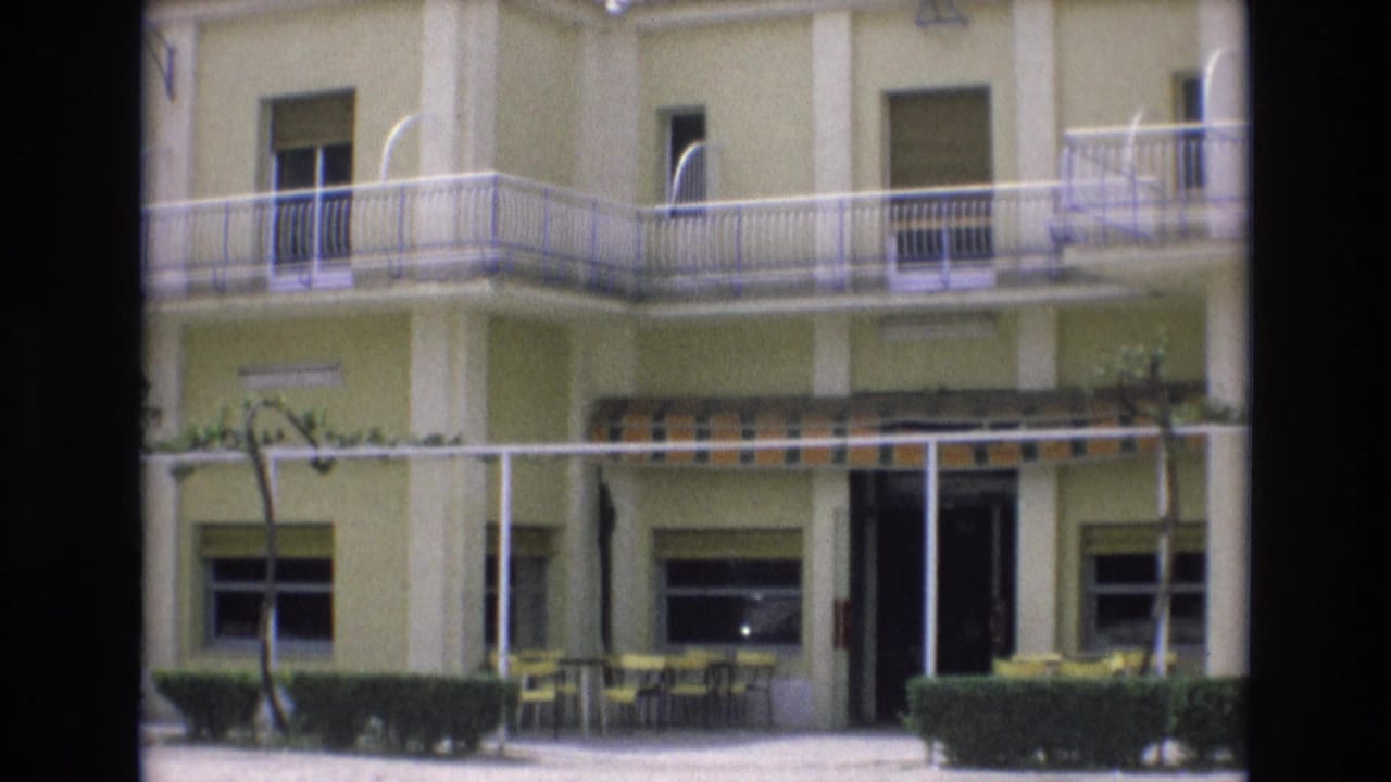 Two-story motel facade with balcony railing, striped awning, patio seating, hedges, and open doorway.