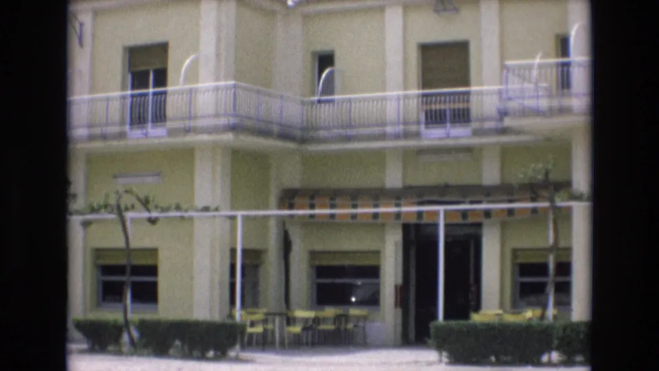 1972 Spain frame showing the motel facade from outside, with balcony railings, shuttered windows, striped awning, yellow patio furniture, hedges, and an open ground-floor doorway.