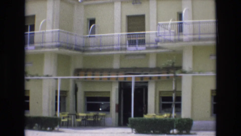 1972 Spain home-movie frame with the motel entrance area, upper balcony, pale facade, striped awning, patio chairs and tables, hedges, and a dark doorway.