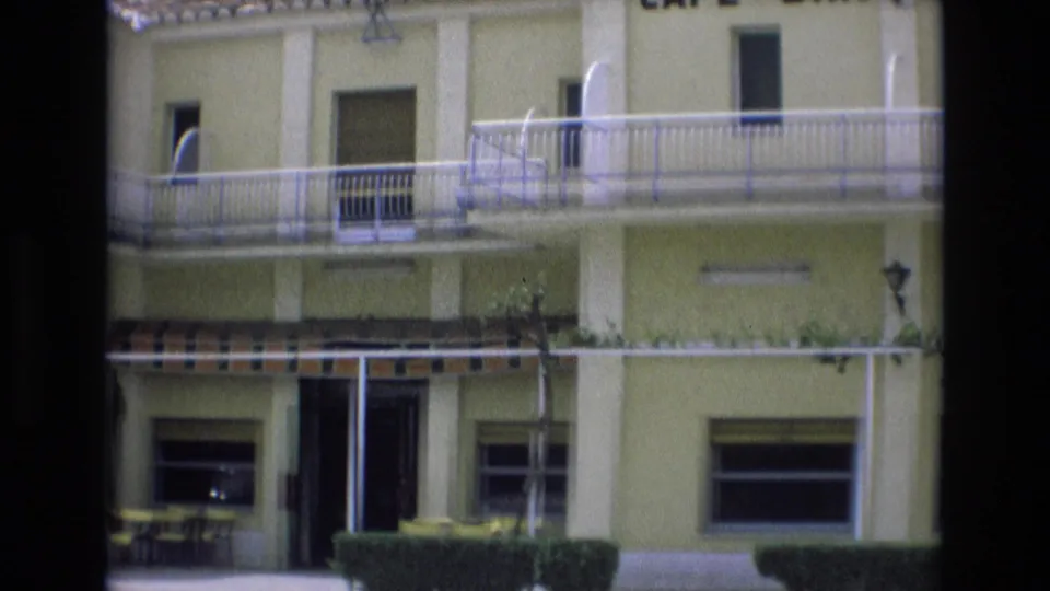 1972 Spain frame showing the pale motel exterior with upper balcony, white railings, striped awning, ground-floor windows, patio furniture, hedges, and dark 8mm frame borders.