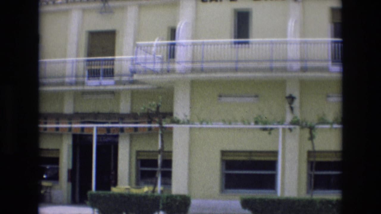 1972 Spain 8mm frame showing the lower and middle facade of a pale yellow cafe-restaurant building with white balcony railings, shuttered windows, striped awning, and trimmed greenery.