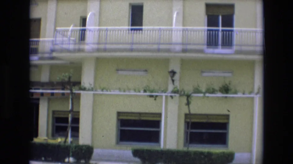 1972 Spain 8mm frame of the cafe-restaurant exterior, with white balcony rails, shuttered windows, ground-level awning, small shrubs, and no visible people.