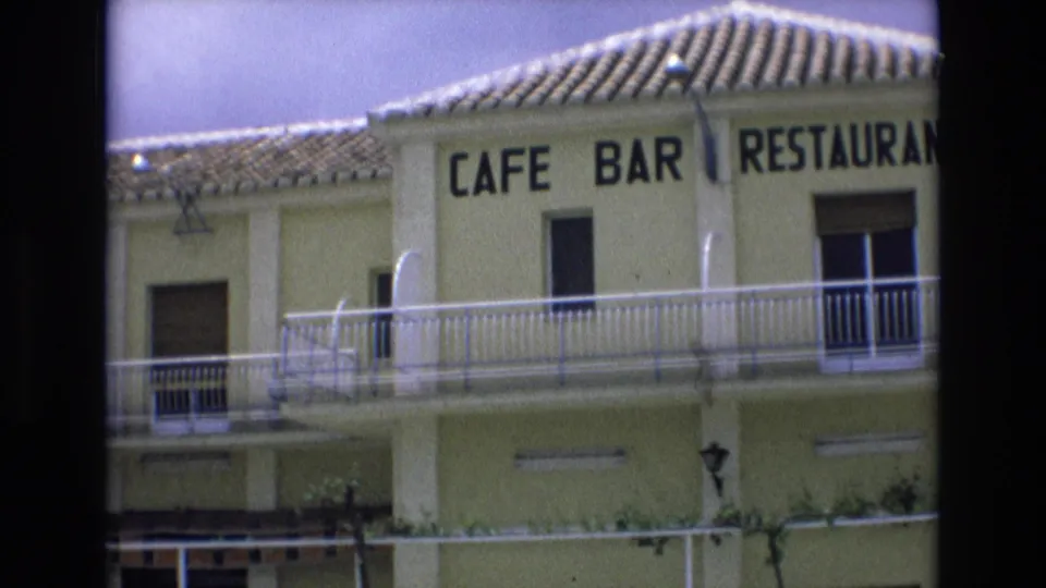 1972 Spain 8mm frame showing the upper facade of a yellow building with a tiled roof, white balcony railings, and the readable CAFE BAR RESTAURANTE sign partly across the wall.
