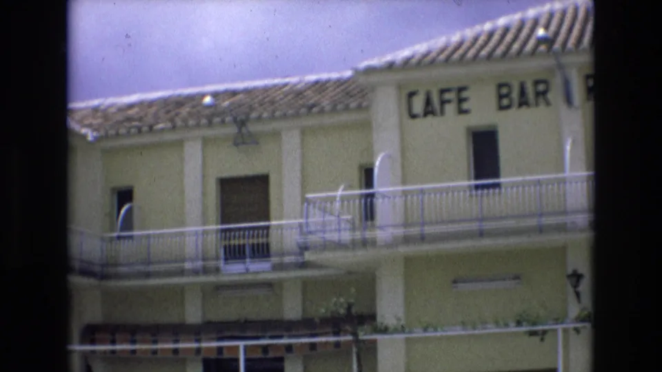 1972 Spain 8mm frame showing a partial CAFE BAR sign on the yellow building, with tiled roof, balcony railings, shuttered openings, and a striped awning below.