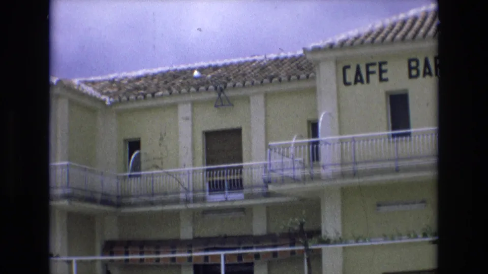 Spain 1972 home-movie frame of a pale yellow two-story cafe bar building with tiled roof, balcony railings, shuttered windows, and partial CAFE BAR lettering.