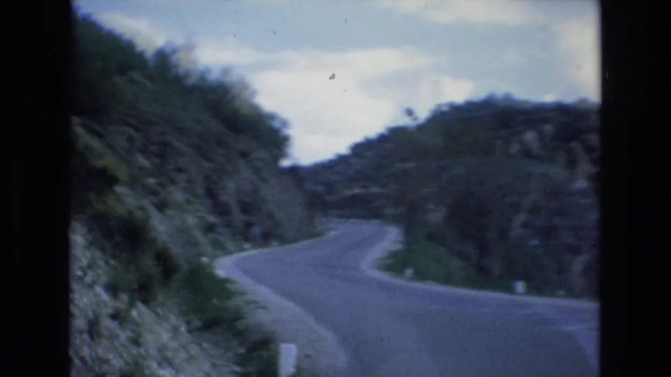 1972 Spain 8mm frame of a narrow mountain road bending through steep, scrub-covered slopes under a partly cloudy sky.