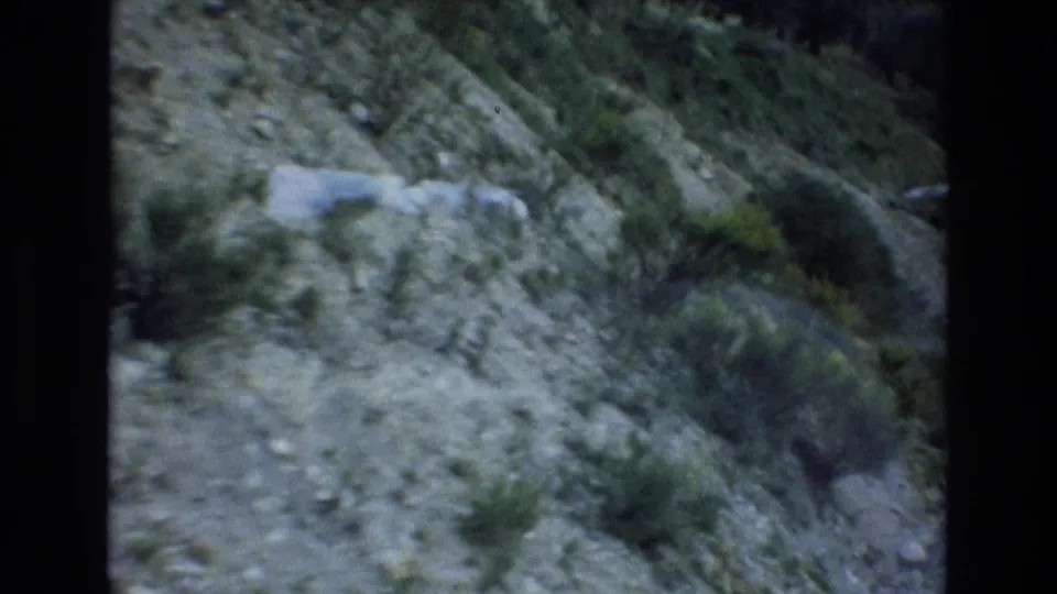 Close 1972 Spain home-movie view of a rocky roadside embankment with low vegetation as the camera passes along the mountain road.