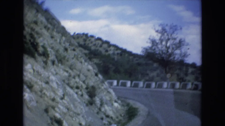 1972 Spain home-movie frame showing a mountain road curving past a white roadside barricade, rocky slopes, scrub vegetation, and a bare-branched tree.