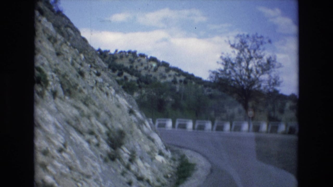 1972 Spain 8mm frame showing a paved road bending around a rocky mountainside, with a concrete barrier and sparse hillside vegetation under a cloudy sky.
