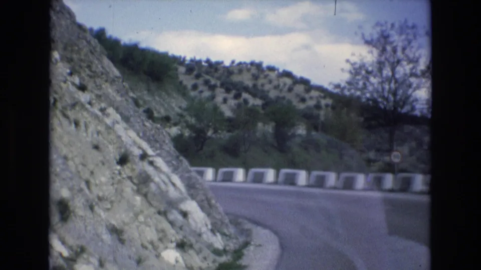 1972 Spain 8mm frame showing the vehicle approaching a bend on a mountain road, with a rocky slope at left and sparse vegetation beyond the barrier.