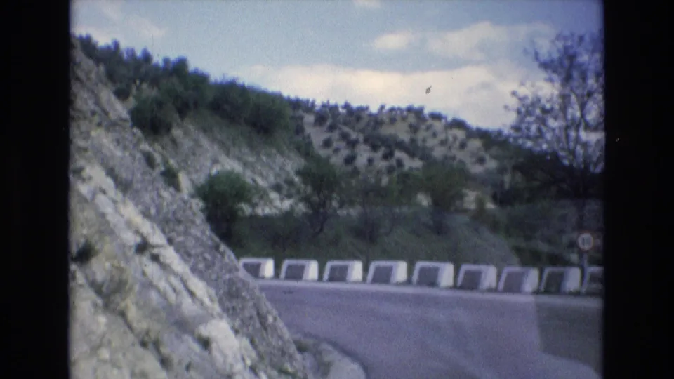 1972 Spain 8mm frame showing a road curve from a moving car, with concrete barrier blocks, a rocky mountainside, scattered shrubs, and trees on the arid hillside.