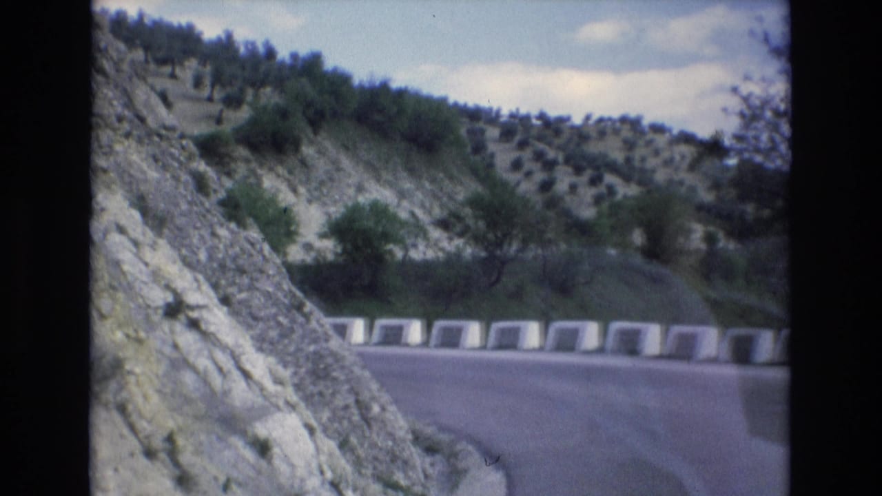 1972 Spain 8mm frame showing a curving asphalt road bordered by white concrete barriers, with a rocky embankment and terraced hillside with small trees beyond.