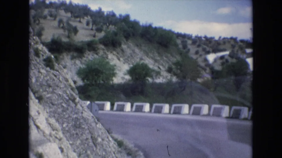 1972 Spain 8mm frame looking along a hillside road curve with white roadside barriers, exposed pale rock, sparse greenery, and small trees on the slope.