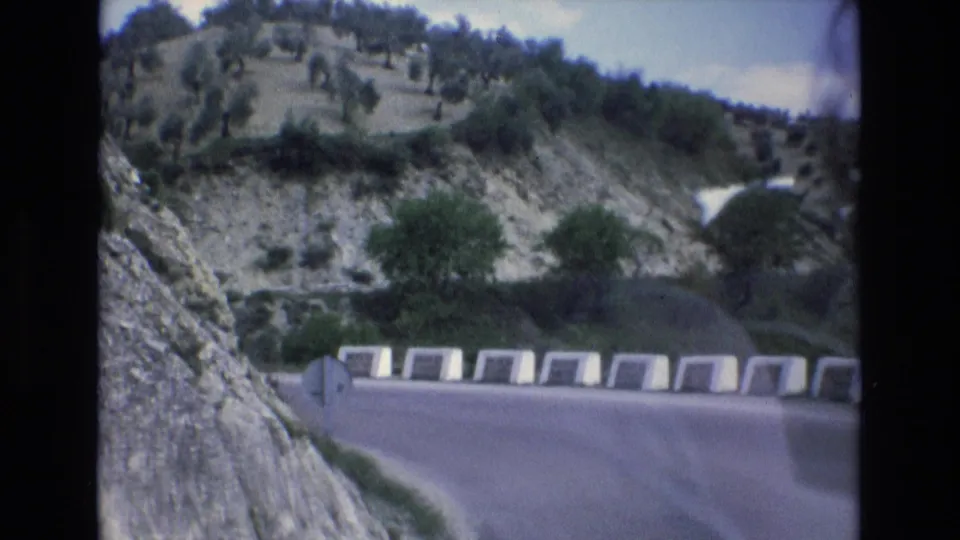 1972 Spain 8mm frame showing a roadside sign near a curving paved road, with white barriers, a rocky embankment, and terraced hillside vegetation in view.
